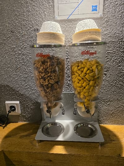 Two cereal dispensers are attached to a wall, one above the other. The top dispenser contains yellow cereal, possibly corn flakes, and the bottom one holds granola. Clear plastic containers display the cereal. Each dispenser has a stack of paper bowls below it. A silver-colored metal frame holds the containers and has two small bowls attached to the side. A power outlet is visible on the wall below the bottom dispenser. The wall behind the dispensers is gray, while the wall to the right is a warm brown wood.