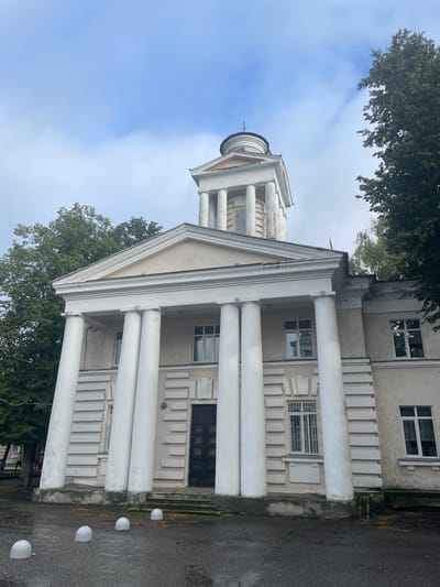 An imposing, classical-style building with a prominent portico featuring large white columns stands against a cloudy sky. The building's facade is light-colored, with decorative rectangular panels and large windows. A dark, double door is visible at the center of the main entrance. To the right and left of the building, tall, leafy green trees are present. In the foreground, a wet, asphalted area is dotted with several round, concrete bollards. The scene appears to be set in an urban environment in Zavolzhye, Russia.