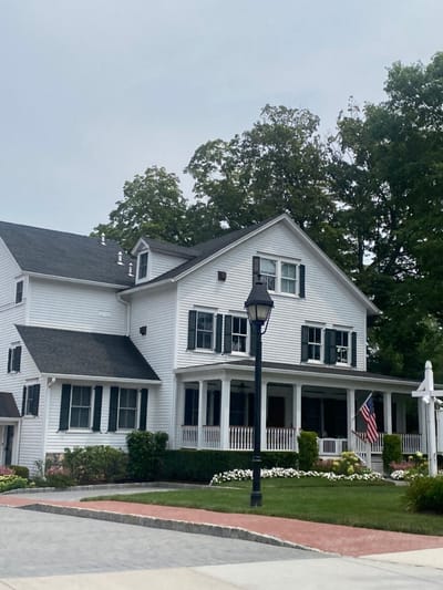 The image displays a large, multi-story white clapboard building with dark grey shingled roofs and dark green window shutters, situated in Ridgefield, United States, under an overcast sky. A prominent front porch, supported by white columns with matching balustrade railings, extends across the facade. An American flag is mounted vertically on the porch railing to the right. A black, ornate streetlamp stands on a manicured green lawn in the mid-ground. Landscaping includes trimmed hedges, shrubs, and flowering plants with white blossoms bordering the foundation and porch. In the immediate foreground, a light grey paved surface is edged by a red brick curb and an adjacent red-brick path. No human subjects or discernible activity are present.