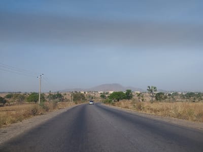 A paved road stretches into the distance, flanked by dry, brown vegetation and scattered trees. A lone car travels towards the viewer, its headlights barely visible. In the background, a small town, likely Taiyere, Nigeria (latitude: 9.83, longitude: 11.364), appears nestled at the foot of a low-lying hill. Overhead, a clear blue sky with a few wisps of white clouds dominates the scene. Power lines and poles mark the roadside, adding an industrial element to the otherwise natural setting.
