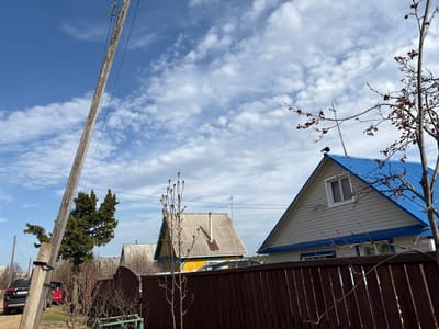 The image captures a rural or suburban residential scene under a clear sky with scattered white clouds. The perspective is slightly upwards, looking over fences and houses towards the horizon.
In the foreground, on the left, bare tree branches with some dried, reddish leaves are visible. To the right of these branches, the gable end of a house with light-colored siding and a bright blue corrugated metal roof is prominent. A single window is centered on this gable. Above the window and partially obscured by a dark wooden fence, Cyrillic text reading "КВАРТИРЫ" (Apartments) and "371" is visible on the house's exterior. The fence, made of dark brown wooden planks, runs horizontally across the upper-left portion of the image, appearing to separate properties.
Further into the scene, another house with a lighter-colored corrugated metal roof (possibly white or light yellow) and greyish siding is seen. A small, dark, shovel-like object or antenna is mounted on its roof. More bare tree branches with small buds or dried leaves are in front of this house.
Towards the right side of the image, a large, dark green coniferous tree stands. A sturdy wooden utility pole, equipped with insulators and power lines, extends vertically upwards on the far right. Behind the tree and pole, parts of a red car and a black SUV are visible. The black SUV's license plate displays "T083BB" and region code "138".
The weather appears to be bright and clear daytime, with blue sky dominating the upper right portion and transitioning to a lighter blue with white, puffy clouds towards the left. There is no specific activity taking place; the scene depicts a static outdoor environment.