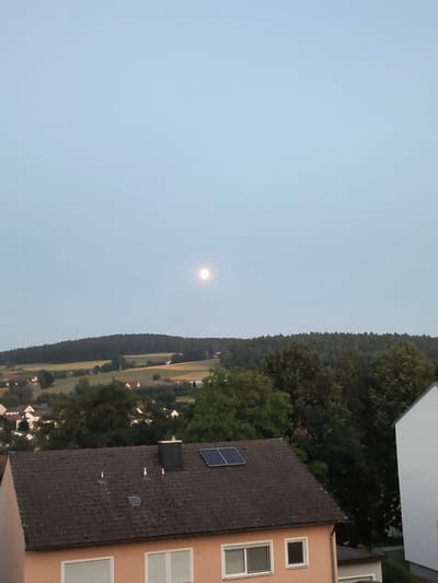 The image depicts a hazy, twilight sky over the city of Oberviechtach, Germany. A full moon is visible in the upper center of the frame. In the foreground, a residential building with a brown tiled roof is partially visible. Solar panels are mounted on the roof. The building has orange-colored walls and two windows with white frames. Beyond the building, a treeline forms a dark, undulating silhouette against the lighter sky. Further in the distance, rolling hills with scattered houses and fields are visible. The overall atmosphere is serene and quiet, characteristic of dusk in a rural or semi-rural setting.