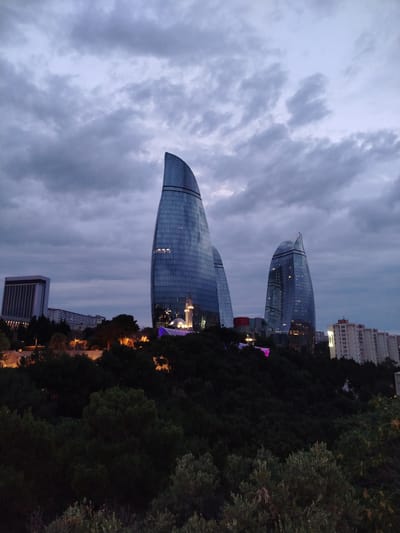 The image shows the Flame Towers in Baku, Azerbaijan, at dusk. The three towers, shaped like flames, are illuminated and reflect the twilight sky. In the foreground, there is a dark, tree-covered hillside. In the background, other buildings and city lights are visible. The sky is cloudy and overcast, with a bluish-gray hue. The overall impression is one of modern architecture set against a natural landscape, with the city coming to life as evening falls. The Flame Towers are a prominent landmark in Baku, symbolizing the city's oil and gas industry.