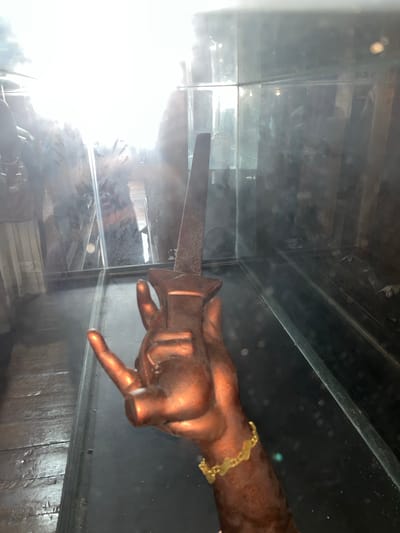 A copper-colored hand sculpture, holding a metallic dagger, is positioned in the foreground. The sculpture is within a glass enclosure that reflects its surroundings, including what appears to be a wooden floor and veiled figures in the background. A gold bracelet adorns the wrist of the sculpture. The scene is brightly lit from the left, causing significant glare and obscuring details in that area. The location is described as Mesjidpirak, Indonesia.