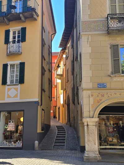 The image shows a narrow, sun-drenched street in Bellinzona, Switzerland, lined with colorful buildings. The street appears to be a pedestrian walkway, paved with cobblestones, and curves gently to the right, leading upwards towards a set of stone stairs.
On the left side of the street, buildings feature balconies with decorative iron railings and windows adorned with green shutters. The facades display a mix of architectural styles, with some sections painted in soft yellow and others featuring decorative stucco work. A street sign reading "PIAZZA NOSETTO" is visible on one of the buildings.
On the right side, a shop window displays mannequins dressed in clothing, with signs advertising "SALDI" (sales) offering 30% and 50% discounts. The shop appears to be a clothing boutique. Above the shop, an archway with intricate decorative patterns leads into a building.
The time of day is clearly daytime, with strong sunlight casting shadows and illuminating the scene, suggesting fair weather. The overall impression is one of a charming and lively European street, likely in a historic town center, bustling with commercial activity and architectural beauty. The image is oriented vertically, with the sky visible at the top and the street extending downwards.