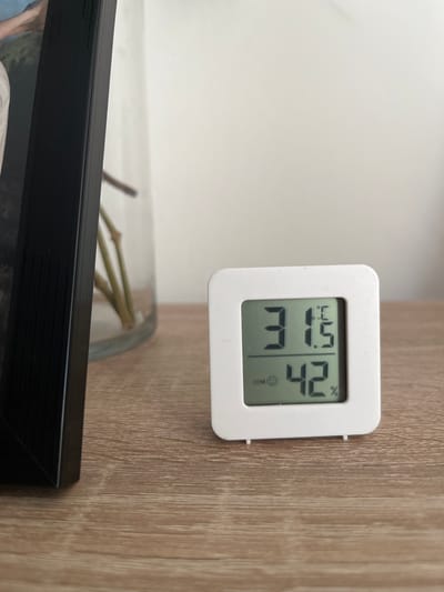 The image shows a white digital thermometer and hygrometer on a wooden surface. The device displays a temperature of 31.5 degrees Celsius and a humidity of 42%. To the left of the device is a black picture frame, and behind it, a glass vase containing several plant stems is partially visible. The background is a plain, light-colored wall. The scene appears to be an indoor setting, likely a room in a home or office. No specific location in Břeclav, Czechia is indicated.