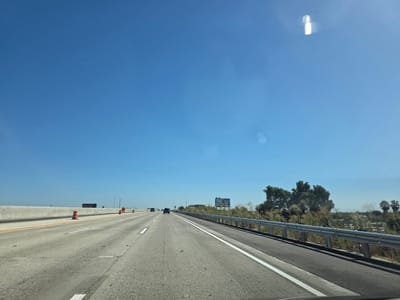 The media file depicts a view from a vehicle traveling on a highway under a clear, bright blue sky. The highway has multiple lanes, and traffic appears light with a few cars visible in the distance. Orange traffic cones are placed along the side of the road, indicating construction or a lane closure. A concrete barrier separates the highway lanes from an adjacent area.
To the right of the highway, there is a metal guardrail bordering a vegetated area with dry grasses and some trees. In the distance, a billboard can be seen with colorful graphics and text, though the specifics are illegible. The time of day appears to be midday, given the bright sunlight and lack of shadows. The weather is clear and sunny.
The scene suggests a typical highway driving experience on a sunny day in a potentially rural or suburban area, given the roadside vegetation and open sky. There is no discernible emotion conveyed. The location is identified as West Sacramento, United States of America.