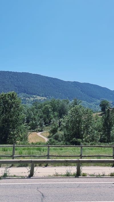 This is a landscape photograph taken outdoors on a clear day, likely in the morning or afternoon given the bright sunlight. The image captures a scenic view of a mountain range covered in dense, dark green forest, transitioning to lighter green grassy slopes lower down. A clear blue sky dominates the left side of the frame.

To the right of the mountain, a line of lush green trees forms a distinct border between the natural landscape and a man-made path. This path, appearing to be made of gravel or dirt, curves gently through a grassy area. Separating the path from a paved road on the far right is a rustic wooden fence. The road itself is asphalt with a white painted line, showing some cracks and wear, suggesting it is a public road.

The overall scene depicts a tranquil setting that balances natural beauty with the presence of human infrastructure, possibly a park or a scenic route in a rural or mountainous area like Llanars, Spain. There are no people or animals visible in the photograph, and no text is present. The image conveys a sense of peace and natural splendor.