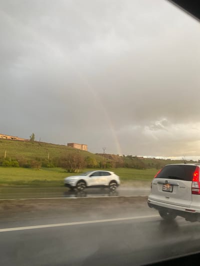 The image depicts a rainy highway in Kotayk, Armenia. A white Honda CR-V is ahead of a silver sedan driving on the road. In the background, a rainbow is visible against an overcast sky over a grassy hillside with buildings. Power lines traverse the landscape.