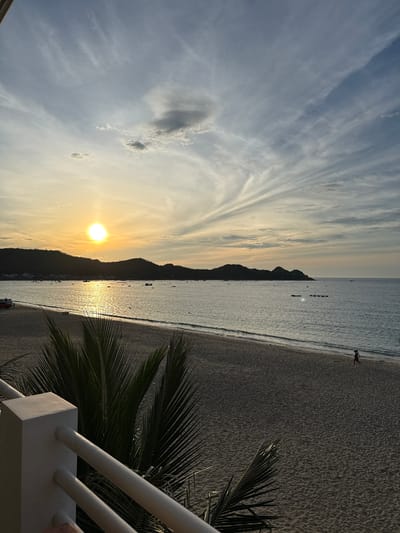 This is a photograph taken from a balcony overlooking a beach in the city of Song Cau, Vietnam, during sunset. The sun, a bright orb, is setting over the ocean, casting a warm, golden glow across the sky and water. Wispy clouds stretch across the sky, painted in hues of orange, yellow, and blue. In the distance, a silhouette of hills forms the backdrop to the scene, with a scattering of small boats visible on the calm sea. The sandy beach stretches along the coastline, with a single figure walking along the water's edge. In the foreground, a railing and the fronds of a palm tree frame the view. The overall atmosphere is peaceful and serene, capturing the beauty of a coastal sunset.