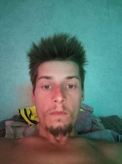 A head-and-shoulders, front-facing selfie features a young, fair-skinned male with spiky dark hair and a thin goatee. He has blue eyes, prominent cheekbones, and a somewhat splotchy complexion with redness around his nose and cheeks. He is bare-chested. In the background, to his right, are several folded gray and yellow fabric items, possibly towels or blankets, leaning against a teal-colored wall. The lighting is dim and cast from the left, creating a reddish-pink hue on the right side of his face. The image appears to have been taken indoors. There is no information available about the location being in Zīlāni, Latvia.