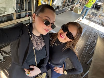 Two women in sunglasses pose for a selfie inside what appears to be an old aircraft. The woman on the left wears a black turtleneck, jeans, and a watch. The woman on the right wears a lace top, a black blazer, and large rings and a gold chain. The interior of the aircraft is visible, with metal walls, windows, and various pieces of equipment. A man wearing a yellow shirt is seen in the background.