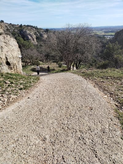 A gravel path descends a hillside. Two people walk away on the path, flanked by trees and grassy areas. To the left is a tall rock face with vegetation at its base. In the distance, a wide landscape is visible under a blue sky with light clouds. The path dominates the foreground, filling most of the lower frame. The scene suggests a natural, slightly rugged environment.