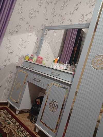 In the city of Karavan, Kyrgyzstan, a dresser with a mirror is pictured. The dresser is light gray with gold accents on its drawers and doors. The mirror reflects a lilac curtain and a wall with floral wallpaper. On top of the dresser, there are several bottles, including Coca-Cola, and a white basket containing various cosmetic items. To the right of the dresser, part of a large wardrobe is visible, also featuring gray doors with vertical ribbed detailing and gold accents. A black trash bag is situated on the floor beneath the dresser. The floor is covered by a rug with a dark red border and a beige patterned center.