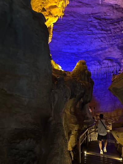 The image shows the interior of the Tenglong Cave in Sanguansi, China. The cave walls are illuminated with blue and yellow lights, highlighting the rock formations. A wooden walkway with railings is visible, and at least two people are walking on it. The foreground is dominated by dark, rocky surfaces.
