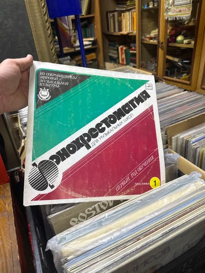 A person holds a vinyl record with Russian text, "Фонохрестоматия для музыкальных школ" (Phonochrestomathy for Music Schools), divided into green and red sections. The record is labeled "Первый Год Обучения" (First Year of Study) and "Пластинка 1" (Record 1). It features the "Melodiya" logo. The environment includes a shelf with various objects and a stack of vinyl records in protective sleeves. This image was taken in Nikolo-Beryozovka, Russia.