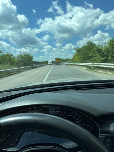 This media file is a first-person perspective shot from inside a car driving on a highway near Sarny, Ukraine. The view looks forward and to the left, capturing the road ahead, a guardrail, and a lush green tree line. The sky is a vibrant blue, dotted with fluffy white clouds, suggesting fair weather. In the distance, other vehicles are visible on the road, including what appears to be a truck.
The car's dashboard is prominently featured on the right side of the frame. It includes a speedometer with markings up to 240, and a tachometer. The time displayed on the dashboard is 13:04. Some text is visible on a small display screen, reading "• В небі, в небі скоро буд..." which translates to "• In the sky, it will soon be in the sky...". This could be a navigation prompt or a message from the car's system. The steering wheel is partially visible in the foreground. The overall impression is of a daytime drive through a scenic, possibly rural, landscape.