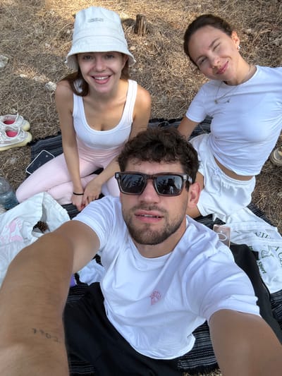 The image depicts three individuals engaged in an outdoor social gathering. The scene takes place in a natural, uncultivated area characterized by dry, light brown ground cover, sparse vegetation, and small rocks. This environment is consistent with a park or open natural space within the city of Modi'in-Maccabim-Re'ut, Israel, potentially on the periphery of a wooded area, as suggested by tree reflections in sunglasses.

A male individual is positioned in the immediate foreground, holding the camera to capture a selfie. He has short, curly brown hair and a light beard, and is wearing dark-framed sunglasses, a white t-shirt with a pink emblem, and black pants. A tattoo is visible on his left forearm. He looks directly at the camera with his mouth slightly open.

To the left and slightly behind him sits a female with light brown hair, wearing a white "JACQUEMUS" bucket hat, a white tank top, and light pink leggings. She is smiling and looking at the camera.

To the right and behind the male sits a second female with dark brown hair pulled back. She is wearing a white t-shirt and white sweatpants, smiling and winking at the camera. She wears a delicate necklace with multiple small pendants.

All three individuals are seated on a dark-colored, possibly patterned, blanket or mat laid directly on the ground.

Several objects are visible on and around the blanket:
*   A clear plastic water bottle is positioned to the left of the blanket.
*   A pair of light-colored athletic shoes with pink accents is placed on the ground next to the blanket on the left.
*   A dark-colored smartphone is on the blanket near the female on the left.
*   A white plastic bag, partly open and containing one or more pinecones, is located on the blanket to the left of the male. Text reading "VILLA" is discernible on the bag.
*   A light-colored smartphone in a clear case is partially visible on the blanket near the female on the right.
*   A light-colored sandal or slip-on shoe is partially visible on the ground to the right of the blanket.
*   A folded white garment is on the blanket near the female on the right.

The overall interaction suggests a relaxed outdoor gathering, with individuals posing for a photograph.