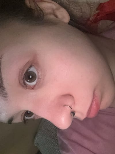 A close-up, angled, low-light photograph shows a person's face, focusing on the eye, nose, and lips. The person has brown eyes and a septum piercing. Their skin appears fair with some redness. The background is blurred and dark, with a hint of pink fabric visible. The location in Olaine, Latvia, is not determinable from the image.