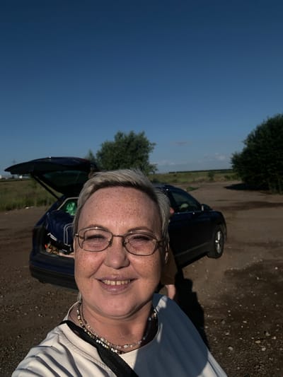 A woman with short, grey hair and glasses is taking a selfie outdoors. She is wearing a white t-shirt and several necklaces. She has freckles on her face and is smiling. Behind her, a dark-colored car is parked with its trunk open, revealing bags inside. The car is on a dirt road, and there are trees and vegetation in the background. The sky is clear and blue. The scene appears to be in Svinogor'e, Russia.