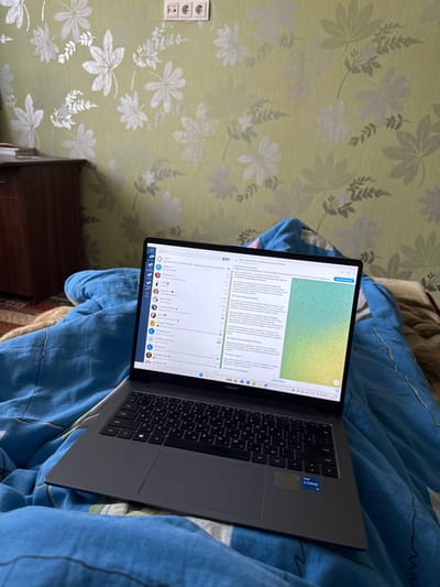 This image depicts an indoor scene in Ciorescu, Moldova, focusing on a dark gray Huawei laptop resting on a blue patterned duvet. The laptop screen is active, displaying a messaging application, specifically Telegram, with a list of contacts and multiple chat windows containing text in Cyrillic script. An "Intel Core" sticker is visible on the laptop's palm rest. Below the laptop, a person's legs or lower body are covered by the blue duvet, indicating the laptop is being used by someone lying in bed.
The background features a light green wall covered with wallpaper adorned with a repeating pattern of silver and white leaves and foliage. To the upper left, a portion of a brown wooden cabinet or chest of drawers is visible. Above the cabinet, mounted on the wallpapered wall, is a white double electrical outlet with two grounded sockets. The overall environment suggests a bedroom or living space.