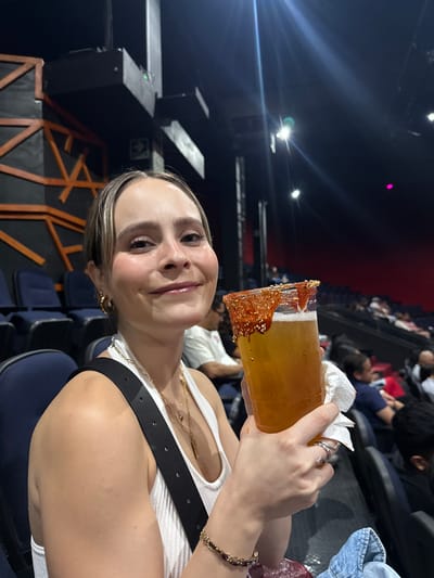 A person holds a tall, rimmed glass filled with a yellow beverage, possibly a cocktail, while sitting in a theater-style seating arrangement. The rim of the glass is decorated with a reddish-orange substance and sesame seeds. The person is wearing a white tank top and a black belt with gold accents and has gold hoop earrings and a bracelet. In the background, rows of seats are visible, some occupied by other individuals. The location is Tlalnepantla, Mexico.