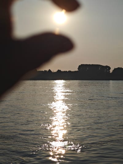 A hand is silhouetted in the foreground, partially obscuring a setting sun. The sun's rays are reflected on the surface of a river, creating a shimmering path of light. In the background, a dark treeline and a small cluster of buildings are visible along the riverbank. The sky is a pale, hazy yellow, suggesting the late hour of the day. The image appears to have been taken from a boat or the edge of the river, looking across the water towards the opposite bank. The scene is peaceful and serene, evoking a sense of quiet contemplation. This scenery is consistent with the natural landscape found in and around the city of Lesosibirsk, Russia, a city situated in the vast expanse of the Siberian taiga, characterized by its numerous rivers and dense forests.