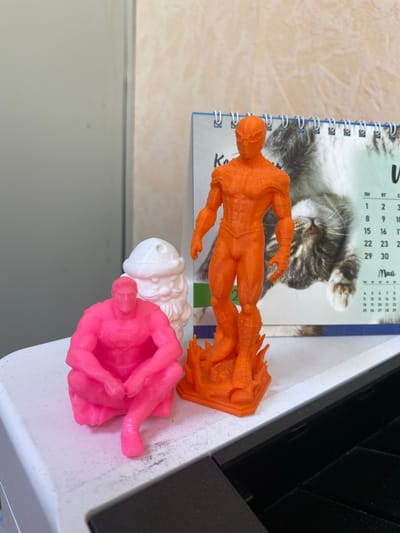 The image shows three 3D-printed figurines: one pink, one white, and one orange. The pink figurine is in a seated, crouched position. The white figurine appears to be a Santa Claus figure. The orange figurine is standing and resembles a superhero. Behind the figurines is a calendar featuring a cat. The figurines are placed on a white surface, possibly a printer or desk, with a dark surface in the foreground. The background is a wall with a textured pattern. The overall scene is indoors. This image does not contain any information about the city of Vyazma, Russia.