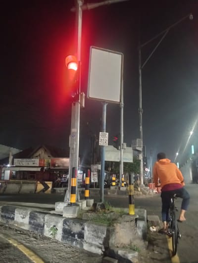 A person wearing an orange hoodie and dark pants rides a bicycle at night in Kedungrejo, Indonesia. The cyclist is seen from behind, facing away from the camera and towards the right side of the frame. The traffic light is red, indicated by a bright red glow. The scene is located on a street with a concrete barrier along the side. In the background, there are buildings with signs, including one that reads "TIARA BAKERY." A tall metal pole with a blank sign is also visible. The sky is dark, suggesting it is nighttime.