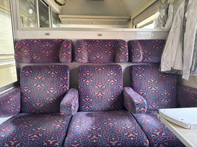 The image displays the interior of a passenger train compartment, located within or operating through Sidi Slimane, Morocco. No individuals are present. The scene is dominated by six upholstered seats, arranged as two rows of three, all covered in a distinctive geometric pattern of blue, red, and white. Each seating unit consists of a lower cushion and an upper backrest. White identification numbers "41", "43", and "45" are affixed to the wall above the corresponding upper backrests. On the left, a window with a curved, decorative metal overlay is visible, reflecting ambient light. On the right side, a white garment, possibly a shirt, enclosed in a clear plastic garment bag, hangs from a hook or rail above the seat. Below this garment, a white, foldable tray table is positioned, partially obscuring the lower cushion of the rightmost seat. The compartment's walls and ceiling are painted a light, neutral color. No actions or interactions are depicted. The environment appears functional and well-maintained.