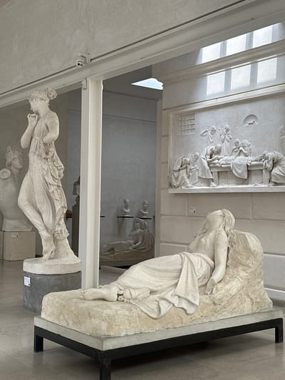 The image depicts a museum interior, likely an art gallery, with several classical-style sculptures. In the foreground, a white marble sculpture of a woman reclining on a stone chaise lounge is prominently displayed. She appears to be asleep or in a state of repose, with her head tilted back and her eyes closed. Her body is draped in flowing robes.
Behind this central figure, to the left, stands another tall white marble statue of a woman in a dynamic pose, her hand raised to her face. To the right of the reclining figure, a large relief sculpture adorns the wall, depicting a scene with multiple figures surrounding a central figure lying down, possibly a depiction of death or a significant event.
In the background, other sculptures are visible, including busts on pedestals and another reclining figure in a more distant alcove. The environment appears to be well-lit, with natural light filtering in from a skylight or a series of windows in the ceiling, casting geometric patterns of light and shadow on the floor and walls. The walls themselves are a neutral, light color. A security camera is visible mounted on a track near the ceiling. The floor is a polished, light-colored stone or concrete. There are no people actively interacting with the sculptures, suggesting the image was taken during a time when the gallery was closed or sparsely populated.
There is no information in the image to suggest the location is in the city of Caniezza, Italy.