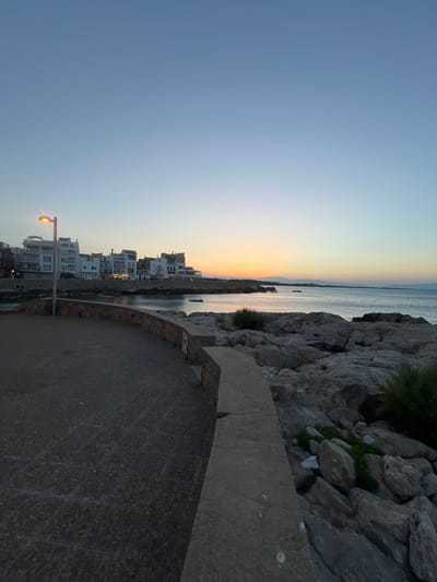 The image depicts a coastal scene in L'Escala, Spain, at dusk. In the foreground, a paved walkway curves towards the left, bordered by a low stone wall. To the right of the walkway, large grey rocks form a natural embankment leading down to the sea. A street lamp with a lit bulb stands on the walkway, casting a warm glow.
Beyond the walkway and rocks, a calm sea stretches towards the horizon. The water reflects the soft colors of the sunset, with hues of orange, pink, and blue dominating the sky. In the middle distance, a line of white buildings with multiple stories lines the coastline, suggesting a town or village. Small boats are visible on the water. The background shows distant hills or mountains silhouetted against the twilight sky. The overall atmosphere is serene and tranquil, characteristic of a coastal town at the end of the day.
