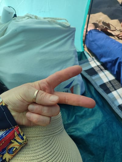 A person's hand, wearing a ring, is held up in a peace sign. The hand is in focus and positioned in the foreground. Behind the hand, there are several objects, including a white cylindrical object with a black cord, a crumpled white fabric, and a folded blue item with a checkered pattern. The scene appears to be inside a tent, suggested by the fabric colors and the presence of what looks like a tent's interior lining. The background shows a glimpse of a sandy area, possibly a beach, with sunglasses visible. This suggests the location is a beach campsite. The image is shot from a close-up perspective, emphasizing the hand and the immediate surroundings within the tent. The overall impression is a casual, outdoor setting, likely a vacation or day trip. There is no direct indication of this location being specifically in Kandıra, Turkey, beyond the prompt's instruction to include this information.