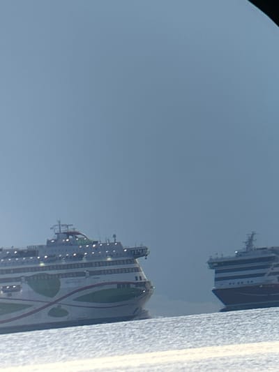 Two large ferries are visible in the water, under a clear, bright sky. The ferry on the left is predominantly white with green and red decorative elements. The ferry on the right is dark blue with white stripes. The water is choppy and appears to be illuminated by sunlight. The scene is likely taking place in Haabneeme alevik, Estonia, given the context of the ferries. There are no people or other objects clearly visible in the image.