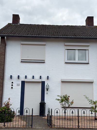 The image shows the facade of a two-story white brick house in Melick, Netherlands. The house has a dark tiled roof with two brick chimneys. The lower level features a closed roll-up door with a dark blue border, flanked by a decorative black mailbox on the right and a bush with flowers on the left. Above the roll-up door, there are three circular openings and a house number "12" on a decorative tile, all set against the white brick. The upper level has two windows. The left window is covered by a roll-up shutter, while the right window has white curtains visible behind the glass. A black metal fence with decorative finials runs across the front of the property, separating the house from the paved walkway and the street. The sky is overcast.