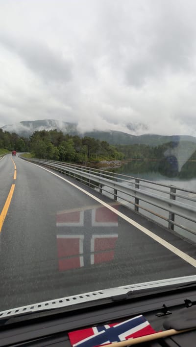 A photograph taken from a vehicle's perspective shows a road with a reflection of the Norwegian flag on its wet surface. The road curves to the left and is bordered by a metal guardrail on the right, separating it from a body of water. The water reflects the surrounding landscape of green trees and misty mountains under a cloudy sky. In the distance, a red truck is visible on the road. A small Norwegian flag is also present on the dashboard of the vehicle in the foreground. The location is identified as Engan, Norway.