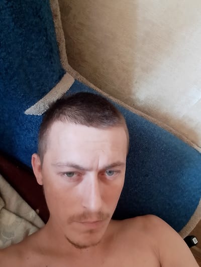 A man with short brown hair and light blue eyes is looking at the camera with a furrowed brow. He has a light mustache and goatee. He is shirtless and appears to be indoors, with a blue carpet and a beige wall visible in the background. The image is taken from a slightly high angle.