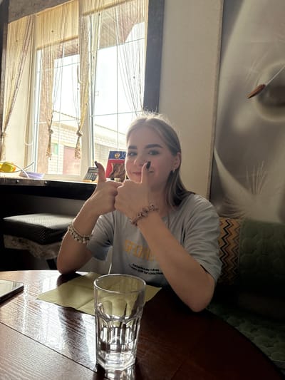 This indoor photograph, taken in Krapivka, Russia, features a young woman looking directly at the camera and giving two thumbs-up gestures, indicating approval or positivity. She has long, light-colored hair and is wearing a light grey t-shirt with partially visible text, including "SFODD," "Collectibles," "d Boxes," "th For," and "Good Frie." She accessorizes with a scrunchie-like bracelet on her left wrist and a white beaded bracelet on her right.
The woman is seated at a wooden table, where a clear glass rests on a light green placemat. Bright natural light floods the scene from a large window to her left, suggesting it is daytime. The window is adorned with intricate string or bead curtains, and a hint of the exterior is visible beyond. A small yellow object can be seen on the windowsill.
The background reveals an interior with varied wall coverings: a subtle textured pattern behind her head transitions to a more pronounced zigzag geometric pattern further down and to the right. A ceiling light fixture with a small decorative pendant is also visible at the top of the frame. The overall atmosphere is casual and cheerful.