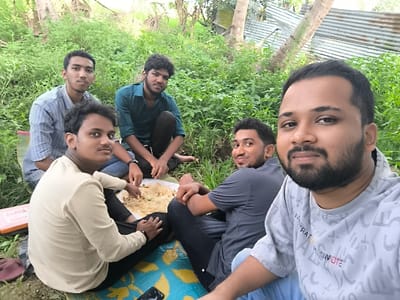 The image shows four young men sitting on a blue and yellow mat, eating rice and vegetables from a white styrofoam plate. The scene takes place in a grassy area near a corrugated metal fence in Kalmunai, Sri Lanka. The men are wearing casual clothing and are sitting with their legs crossed. The man on the right of the image is smiling at the camera. The man in the center of the image is looking to the left. The man on the left of the image is looking at the camera. The man in the background is looking down at the plate. The environment is green and lush, with palm trees in the background. There is a red book on the ground in the bottom left of the image. There is a black cell phone on the mat in front of the men. There is a white notebook on the ground to the left of the men. The image was likely taken on a sunny day.