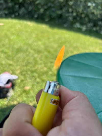 An extreme close-up shot captures a person's hand holding a yellow BIC lighter with a flame. The background is a blurred outdoor scene with green grass and trees. The focus is on the lighter and the flame. There is no information in the image that suggests a location in Vicenza, Italy.