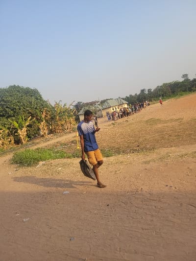 The image depicts an outdoor scene in what is identified as San Martín, Colombia, under a clear, light blue sky. A young adult male, dressed in a blue and white striped short-sleeve shirt and orange shorts, with sandals on his feet, walks from the left to the right side of the frame. He carries a dark, possibly black, bag or sack in his left hand and casts a long shadow to his left. The ground is a reddish-brown dirt or sandy surface with visible tire tracks and scattered small white debris. To the left, dense green foliage, including large trees and what appear to be banana or plantain plants, line the edge of the dirt path. In the middle ground, several light-colored buildings with green or gray sloped roofs are visible. A substantial group of people is gathered in front of these buildings, arranged in what appears to be lines or queues. Further to the right, beyond the gathered crowd, there is an expansive, dry, reddish-brown dirt field with a lone figure in red visible in the far distance, possibly running or walking. Additional trees and vegetation are present on the horizon.
