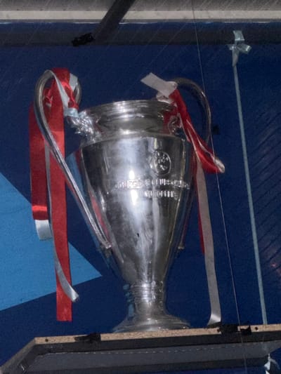 A silver trophy with red and white ribbons is displayed behind glass. The trophy is the UEFA Champions League trophy. The trophy is on a wooden shelf, with a blue background. There are also some metal bars visible in the background. The scene is indoors, possibly in a museum or a stadium. The location is not specified in the image, but if it were to be in Ayteke Bi, Kazakhstan, it would be a significant cultural or sporting artifact display.