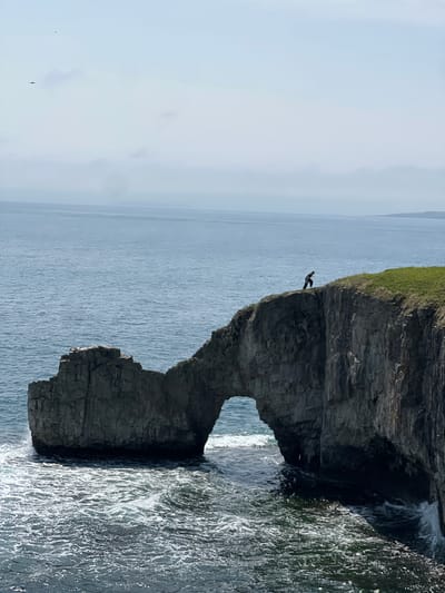 The image captures a coastal scene featuring a dramatic rock formation with a natural archway carved by the sea. The rock is dark grey and textured. A small figure stands on the top of the rock. The sea is visible through the archway, appearing light blue and calm in the distance, with darker, more turbulent water closer to the rock face. The coastline is characterized by a grassy area atop the cliffs. The location is the Bruce Peninsula near Slavyanka, Russia.
