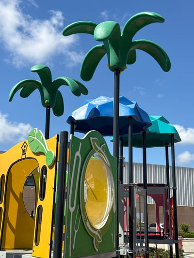 A low-angle shot captures a section of a playground against a bright blue sky with scattered white clouds. The playground equipment is colorful, featuring green, blue, and yellow sections. In the foreground, two green, stylized leaf-like structures with curved blades are visible, extending outwards. Behind these, a large blue, faceted dome-like structure and a similar teal structure are prominent.

Further back, the playground structure includes yellow panels with cut-out shapes and green accents, one of which has a circular window with a yellow interior. A metal frame with red and gray components is visible higher up, suggesting a more complex play structure. Some red elements appear to have the word "TALIYA" and "ALUM" visible on them.

The lighting suggests it is daytime, likely sunny, given the clear sky and bright colors. The overall impression is of a vibrant and modern playground designed to engage children. The scene is static, with no people or specific activity visible, but the design of the equipment implies a place for active play. The presence of trees in the background suggests an outdoor setting, possibly a park or recreational area. The location context is Pardes Hana - Karkur, Israel.