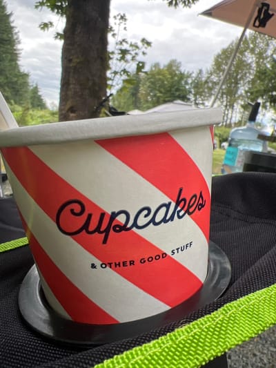 The image shows a close-up of a red and white striped paper cup that says "Cupcakes & Other Good Stuff" in black cursive lettering. The cup is sitting in a black cup holder with a green trim, on top of a black fabric surface. In the background, out of focus, there are trees, a large tree trunk, a cloudy sky, a light-colored tent or canopy, and a blue jug of water. Based on the foliage and trees, the location appears to be outdoors, possibly in a park or campground. The presence of a tent suggests this might be a campsite. Without specific landmarks or contextual information within the image, it is not possible to determine the exact location within Chilliwack, Canada. There are no people visible in the image.