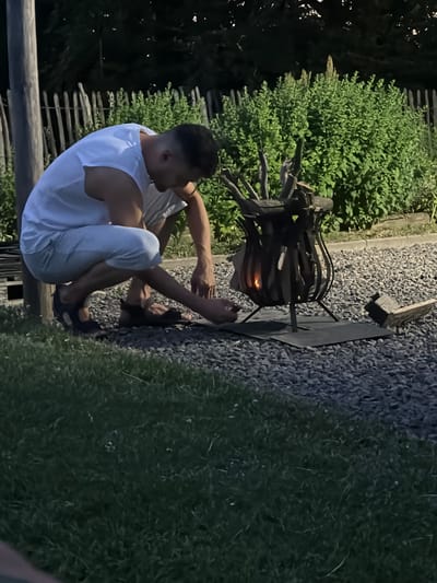 In this image, a man is seated on the ground in what appears to be an outdoor setting at dusk. He is wearing a white tank top and light-colored pants, with his legs bent and tucked towards his body. The man is leaning forward, with his hands near a fire pit. The fire pit is made of metal and contains burning wood, casting a warm glow. The ground around the fire pit is covered in gravel.
Behind the man, there is dense green foliage, likely bushes or trees, and a rustic wooden fence. The overall scene suggests a relaxed, possibly camping or backyard setting. The lighting indicates it is evening, likely sunset or just after, with ambient light fading. There are no visible signs of text in the image. The man's posture and focus on the fire pit suggest he is tending to it or simply enjoying its warmth. The environment appears natural and somewhat wild, given the foliage and rough ground.