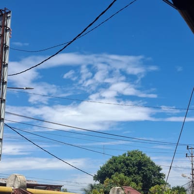 The image captures a bright, sunny day in Mesjid Tuha, Indonesia, dominated by a vivid blue sky dotted with scattered white clouds. The foreground is filled with a dense network of black electrical wires crisscrossing the frame, suggesting an urban or semi-urban environment. On the left side, a tall, rust-colored metal structure, possibly part of a billboard or advertisement frame, stands prominently, with a street lamp attached to it pointing towards the right. Below the wires and partially obscured by them, rooftops of buildings can be seen, one with a reddish-brown tiled roof. To the right, a large, verdant tree with lush green foliage occupies a significant portion of the lower right quadrant, its branches reaching upwards. Further in the background, a thin, tall radio tower is visible against the sky. The overall impression is of an outdoor scene in a populated area, with clear weather conditions.
