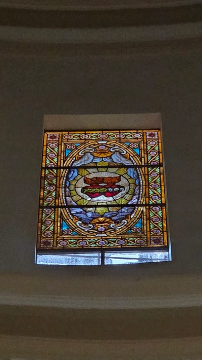 A stained-glass window featuring an oval design with two figures is visible. The window is decorated with colorful geometric patterns and is inset into a wall. Outside, the sky appears bright. The location is the Carthusian Monastery of Valldecrist in Altura, Spain.