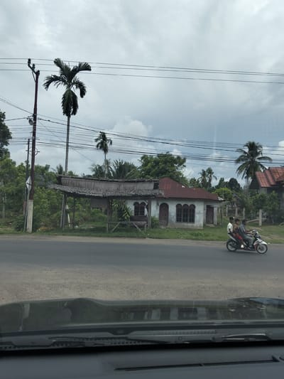 The image depicts a street scene in Meunasahlhok, Indonesia, under a cloudy sky. In the foreground, the hood and windshield of a vehicle are visible, suggesting the photo was taken from inside a car. The main focus of the image is a rural roadside with several structures and natural elements. A paved road runs horizontally across the middle of the frame, with a shoulder of gravel and sparse vegetation.
On the left side of the road, a utility pole with electrical wires is prominent, supporting the overhead lines that stretch across the scene. Behind the pole, lush green trees and foliage create a dense backdrop. A tall palm tree stands out to the left of a rustic open-air shelter with a thatched roof, which appears to be a bus stop or waiting area. This shelter has wooden posts and beams, and a simple bench.
To the right of the shelter, a small building with a red corrugated metal roof and light-colored walls is visible. It has arched windows with dark frames and a traditional architectural style. Further to the right, another building with a similar red roof is partially obscured by trees.
A motorcycle with three people on it is traveling on the road from right to left. The rider is in front, followed by two passengers. The motorcycle is white and red. The people are dressed casually. The overall environment suggests a tropical climate with abundant vegetation and a somewhat overcast day. The scene conveys a sense of everyday life in a rural or semi-urban Indonesian setting.