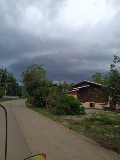 The photograph shows a paved road curving to the left in the city of Pak Phraek, Thailand. The sky is overcast with dark storm clouds. Lush greenery borders the road on both sides. A wooden building with a beige base is visible to the right, near the bend in the road. A motorcycle is parked near the building, where an individual stands. Power lines run overhead.